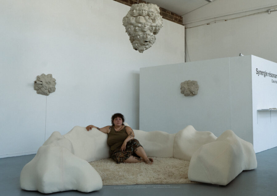 Heller Ausstellungsraum: weiße keramische Objekte hängen an den Wänden und eines von der Decke. In der Mitte eine weiße, gepolsterte Liegelandschaft mit Teppich, auf der eine Person sitzt.
