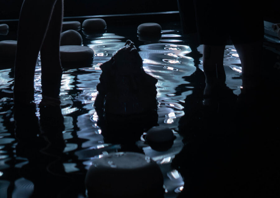 Dunkle Wasserfläche in einem abgedunkelten Raum. Keramische Schwimmkörper treiben darauf, Lichtreflexe spiegeln sich; am Rand Silhouetten stehender Menschen.